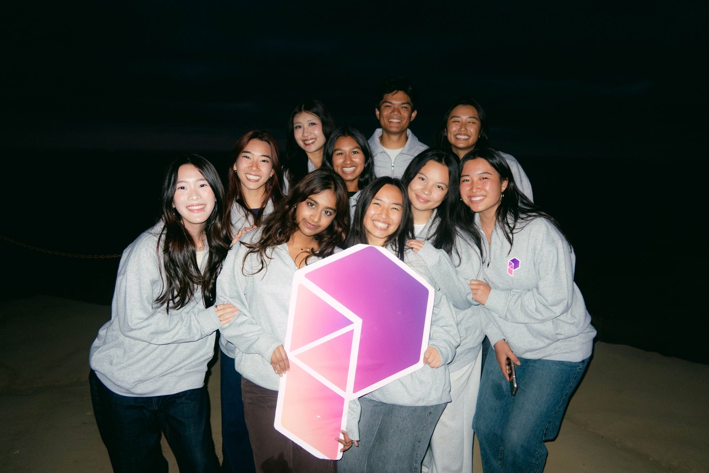 Friends in matching gray sweatshirts on the beach at night, holding a large Product Space logo